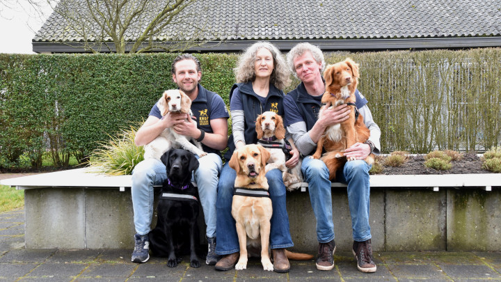 Groepsfoto van vijf medische detectiehonden en hun trainers op een bankje